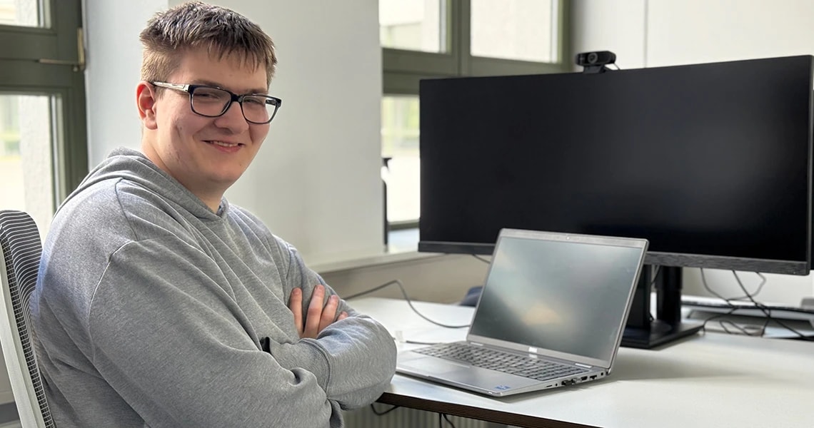 Ivan sitzt am Schreibtisch, Laptop und Bildschirm im Hintergrund